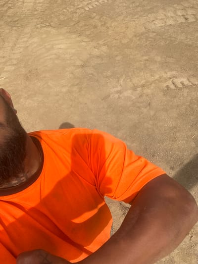 The image, taken in Weissach im Tal, Germany, presents a high-angle view looking downwards, primarily capturing the upper body of a person and the ground beneath them. The subject is a person with dark skin, wearing a bright orange short-sleeved t-shirt and a dark neckband, possibly a collar or a visible base layer, with a faint gold chain. A dark beard is visible on the chin area. The person's right arm and shoulder occupy the right side of the frame, showing a faint tattoo on the inner forearm/bicep region. Both the skin and the orange shirt show some accumulation of dust or dirt.
The setting is outdoors, characterized by strong, direct sunlight casting distinct shadows, indicating a clear, bright daytime. The ground, which makes up the majority of the background, is a sandy or dusty texture, featuring loose soil, small pebbles, and visible tire tracks. These details suggest an unpaved area, such as a construction site, a dirt road, or an open field. The person appears to be sitting or leaning. No specific activity is depicted beyond being present in this environment. There is no visible text in the media file.