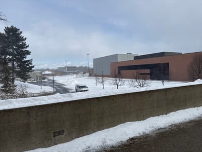Snowy landscape featuring a concrete barrier in the foreground. A road winds through a campus-like setting, with a white van visible. Buildings of varying architectural styles are present, including a modern brick structure with large windows. A tall pine tree stands to the left, and bare trees are scattered throughout the scene. Overcast sky completes the wintry atmosphere.