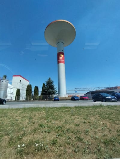 This media file captures an outdoor scene under a clear, bright blue sky, suggesting a sunny day. The perspective is unusual, with the image rotated so that the ground appears on the right side and the sky on the left.
The prominent subject is a tall, cylindrical structure, likely a water tower, dominating the left side of the frame. It is painted white and has a large, reddish-yellow circular logo on its side with the word "Culinar" written in red lettering. Atop the tower is a wide, circular, beige cap.
To the right of the water tower is a paved area with several cars parked along it. A dark blue car, a red car, and a lighter blue car are clearly visible. Beyond the parking area, there is a strip of dry, grassy ground with scattered white wildflowers. A fence runs along the edge of the grassy area.
Further in the background, to the right of the water tower and cars, is a modern building with white walls and a red roof. Several coniferous trees, some tall and slender and others more bushy, are visible in front of the building and along the fence line.
The overall impression is of an industrial or commercial area on a bright, clear day. The image appears to have been taken from a moving vehicle, as evidenced by reflections on a surface (likely a window) in the sky portion of the image. The location is identified as Horažďovice, Czechia. No people are visible in the frame.