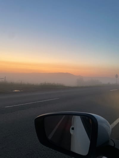 The image depicts a car's side mirror reflecting the interior of the vehicle, including a white door handle and a portion of the car's interior. Outside the car, a road stretches towards a hazy landscape, likely in the city of Uba, Russia, as indicated by the provided context. The scene is bathed in the soft light of dawn or dusk, with a gradient of colors in the sky transitioning from a pale orange near the horizon to a soft blue overhead. A low-lying mist obscures the distant hills and terrain. The road itself is paved and marked with white lines. A few scattered patches of vegetation line the side of the road.