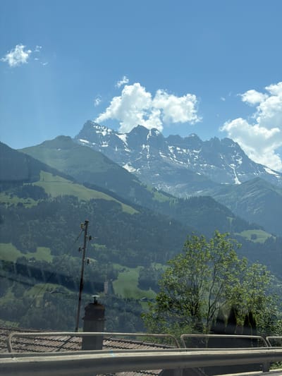 The image depicts a panoramic view of a mountainous landscape, likely in the vicinity of Troistorrents, Switzerland, given the presence of characteristic Swiss Alpine features. The scene is dominated by a majestic mountain range in the background, with its peaks partially covered in snow, suggesting a high altitude and a temperate climate. Lush green slopes, densely covered with trees and interspersed with open fields, descend from the mountains. Some scattered houses or farm buildings are visible on these slopes, adding a touch of rural life to the scene.

In the mid-ground, a prominent wooden utility pole with wires stretches across the frame, indicating human infrastructure in this otherwise natural setting. A chimney, possibly part of a residential building, is visible near the base of the pole. A mature tree with dense green foliage stands to the right of the utility pole, its branches extending upwards and outwards, partially obscuring the view of the landscape behind it.

The foreground is partially occupied by a tiled roof and a metal railing, suggesting the image was taken from a vantage point, perhaps a balcony, terrace, or a moving vehicle. The sky above is a vibrant blue, dotted with fluffy white clouds, which contribute to the overall serene and picturesque atmosphere. The lighting suggests it is daytime, likely late morning or early afternoon, with the sun casting a gentle illumination on the scene. The overall impression is one of natural beauty, tranquility, and the harmonious coexistence of human settlements with the grandeur of the Alps.