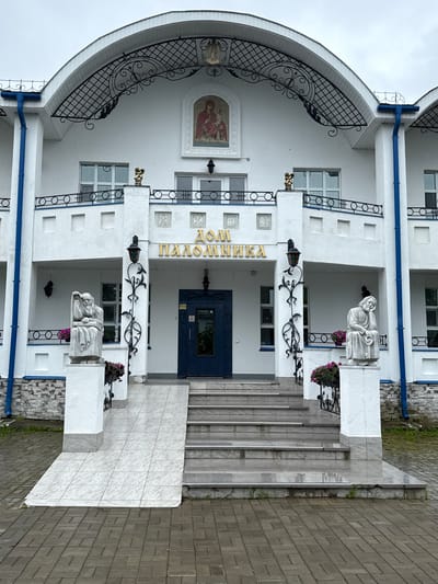 The image shows the white, two-story facade of a building, likely a religious or cultural center, located in Местечко Раифа, Russia. The building features a large, arched metal canopy over the entrance, decorated with intricate scrollwork. Above the entrance, a religious mosaic depicting a woman with a child is displayed. The building's facade is adorned with two sculptures of seated, bearded men, positioned on pedestals on either side of the entrance. Both sculptures are in a contemplative pose. The entrance itself is marked by a dark blue door, surrounded by a set of tiled stairs and a wheelchair ramp. Flower pots with purple flowers are placed near the sculptures. The building is accented with blue trim and blue downspouts. The ground in front of the building is paved with interlocking gray bricks, and the area appears to be slightly wet, suggesting recent rain. The sky is overcast.