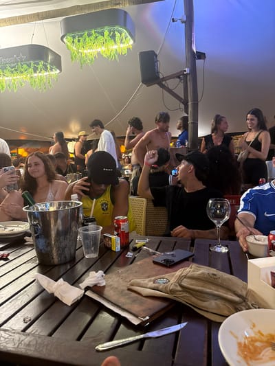 A crowded, semi-enclosed restaurant or bar scene in Cairu, Brazil, under a large white tent or canopy during the evening. Overhead, rectangular light fixtures feature artificial green foliage, with one displaying a "Heineken" logo. A wooden pole supports a black speaker and a white security camera.

In the foreground, a dark wooden slatted table is laden with various items. A silver ice bucket contains a green bottle and two other bottles. Other objects include two red soda cans, a yellow soda can, multiple clear plastic cups, a clear wine glass, a pack of Camel cigarettes, a yellow lighter, loose cigarette filters, a black smartphone, a beige fanny pack, crumpled white napkins, a metal knife, a menu folder, and a white bowl containing food remnants.

Three individuals are seated at the table in the mid-ground. On the left, a woman with dark hair, wearing a white halter top, smiles. Next to her, a male in a yellow Brazil national football team jersey and a black "Vans" baseball cap holds a mobile phone to his ear, looking downwards. To his right, a male in a black t-shirt and black baseball cap makes a gesture with his raised right fist. Further to the right, a male in a blue t-shirt with a white Nike logo is partially visible.

The background shows numerous other individuals, predominantly young adults, standing and sitting, engaged in conversations. Some men in the background are shirtless. The overall scene depicts a lively social gathering.