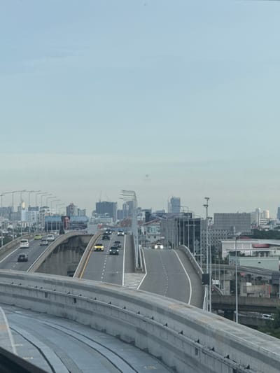 The image depicts an elevated highway system in Pak Kret City Municipality, Thailand. Multiple lanes of traffic are visible on the highway, with various vehicles, including cars and what appears to be a truck, in motion. The highway curves and splits, with some sections appearing to be under construction or newly built. In the background, a city skyline with numerous buildings of varying heights is present, suggesting a densely populated urban environment. The sky is overcast with a light blue hue, indicating it may be early morning or late afternoon. There are no visible individuals in the image. The foreground shows a concrete barrier and what looks like a rail system or another elevated structure. The overall scene captures a snapshot of urban transportation infrastructure within a city.