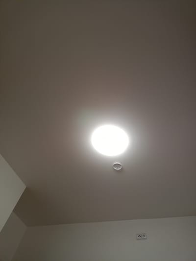 This media file captures an indoor ceiling with a bright circular light fixture illuminating the space. Below the light, a small, circular smoke detector is visible, mounted on the ceiling. Further down, towards the bottom of the frame, a white electrical outlet with two sockets is mounted on a wall. The overall scene suggests a modern interior, possibly residential or commercial, with a focus on functional elements. The lighting indicates artificial illumination, likely from the central fixture, and the absence of windows or external views limits specific environmental cues. The setting appears to be a well-lit indoor environment in Nová Ves nad Lužnicí, Czechia. There is no visible text in the image.