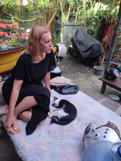 In Bedulu, Indonesia, a fair-skinned woman with blonde hair, wearing a black dress, sits barefoot on a colorful, shaggy rug. Beside her, a black and white cat is curled up and sleeping. A black handbag and several motorcycle helmets are scattered on the rug. A covered motorcycle is parked in the background. The scene is set outdoors, surrounded by lush tropical greenery and potted plants. The woman is looking off to the side, apparently deep in thought.