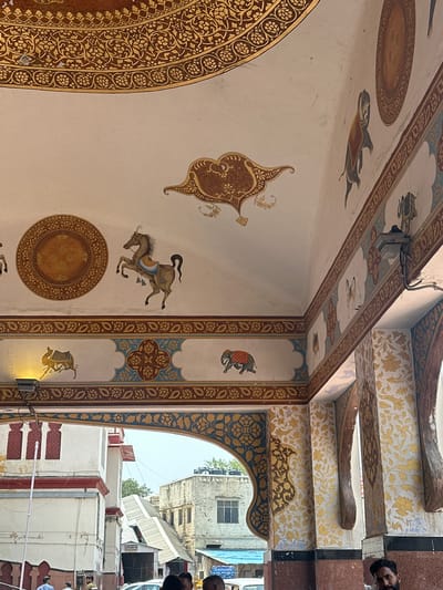 The image shows a close-up view of ornate ceiling and wall paintings within an arched entryway. The ceiling features elaborate golden floral patterns on a brown background, with smaller decorative elements painted on the white surface, including a circular motif and a stylized bird. The walls are adorned with friezes containing painted scenes of animals, such as horses and elephants, interspersed with decorative borders. One section of the wall displays a repeated floral pattern in yellow and white. The entryway opens to an outdoor scene where buildings and some individuals are visible in the background. The style of the artwork suggests a historical or religious site, likely in Ajmer, India, given the cultural context of such elaborate decorative art.