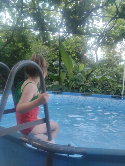 A young girl with brown hair, facing away from the camera, sits on the edge of a blue inflatable pool, holding onto the pool ladder with her right hand. She is wearing a red swimsuit with black watermelon-like seeds and a green rucksack on her back. The pool is filled with clear blue water. The background is lush with green foliage, suggesting an outdoor setting, possibly in Tamarisi, Georgia, where a vine-covered structure with supporting poles creates a canopy overhead.