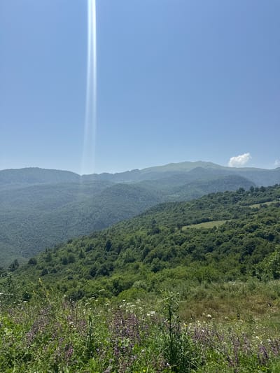 A wide-angle, eye-level, outdoor photograph captures a verdant mountain landscape under a clear blue sky. The foreground is dominated by a field of wild flowers, featuring purple and white blossoms interspersed with tall green grasses and stalks. Beyond this meadow, the middle ground presents rolling hills covered in dense, lush green forests, with patches of lighter green indicating clearings or less dense tree cover. In the background, layered mountain ranges recede into the distance, fading in color to a soft blue due to atmospheric perspective. The sky is a pale blue, with a distinct, bright shaft of light, possibly sunlight or a lens flare, appearing as two parallel white beams near the top center of the image. There are no people, animals, or man-made structures visible in the frame, creating a natural and serene scene. The location is identified as Yenokavan, Armenia.