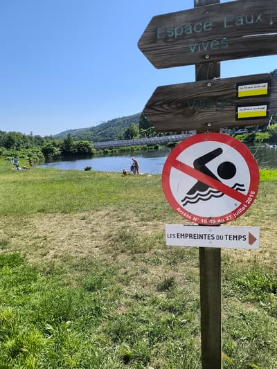 Here's a detailed description of the image:

The scene captures an outdoor setting, likely near a river or body of water, in Saint-Pierre-de-Bœuf, France. The composition is dominated by a wooden signpost with multiple direction signs. The top sign directs towards "Espace Eaux Vives" and another to "Village" with a yellow Le Rhône aménagé logo. 

Attached to the signpost is a circular red and white sign indicating that swimming is prohibited, marked by a swimmer icon crossed out with a red line. Below that sign is a rectangular sign pointing to "LES EMPREINTES DU TEMPS" (The Footprints of Time).

In the background, people are visible near the riverbank, suggesting they might be engaging in activities like sunbathing or wading. A bridge is visible spanning the river in the middle distance. The landscape includes grassy areas, trees, and rolling hills under a clear blue sky. The photo appears to be taken during a sunny day.