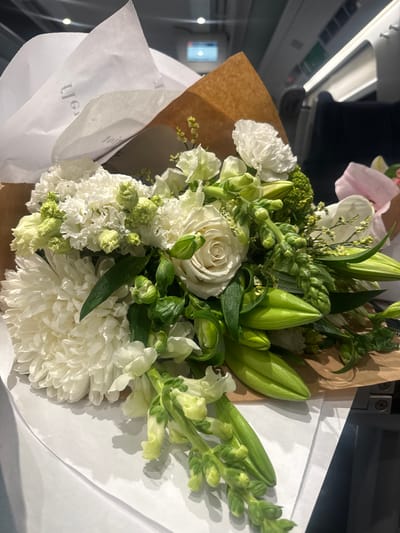 A bouquet of flowers is the central subject of the image. It is composed of white chrysanthemums, lilies in bud, white roses, and other green foliage. The bouquet is wrapped in brown paper and white tissue paper, with text visible on the tissue paper. The flowers are placed on a surface, likely a table or counter. In the background, a partial view of what appears to be the interior of a train or bus is visible. The environment is somewhat blurry, but hints of overhead lights, seating, and windows can be discerned. There is no indication that this image was taken in Domodossola, Italy.