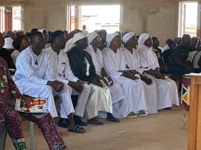 The image shows a group of people, mostly men, sitting in rows indoors. They are all wearing white attire, with some individuals wearing headwraps. The environment appears to be a community hall or a church, with bare concrete walls and a dirt floor. Sunlight streams in from an open doorway or window on the right side of the image. In the foreground, a person is holding a booklet with a picture of a man in a religious garment. To the right, a drum with colorful patterns is visible. The people are looking forward, suggesting they are attending an event or a service. The scene is likely in Kufana, Nigeria, given the context.