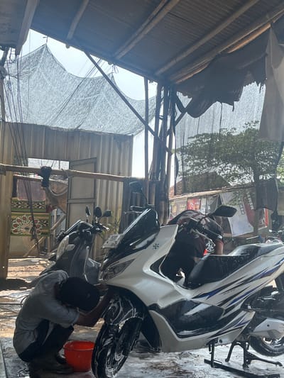 A man washes a white Honda motorcycle at a business in Curug, Indonesia. He crouches next to the motorcycle, which is angled towards the right and covered in soap. A red bucket is next to him. Two other motorcycles are visible in the background. The scene is under a makeshift shelter of corrugated metal, bamboo, and netting. Commercial signs are visible in the background.