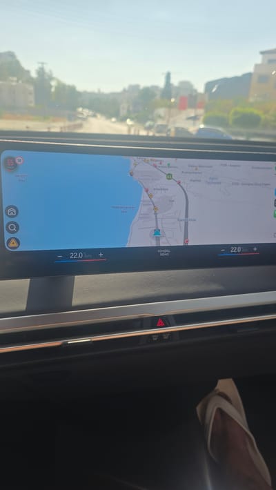 The image shows a car's dashboard display, rotated 90 degrees clockwise. The screen displays a map of an area, likely a coastal region, with roads, buildings, and the Mediterranean Sea labeled. The map shows street names such as "Voreiou peirou," "Steliou Mavromati," and "Andrea Kariou," as well as locations like "Amathus Archaeological Site" and "Akrotiri Bay." The temperature is displayed as 22.0 degrees Celsius, with "AUTO" and "MENU" options visible, suggesting climate control settings. There's also a speed display showing 55 km/h and 50 km/h.
On the right side of the dashboard, a person's hand is visible, holding a light-colored cloth and appearing to wipe something. A hazard triangle warning light is also visible on the dashboard. The overall scene suggests the car is in motion, with the exterior view visible through the windshield showing buildings and trees under a bright, possibly sunny sky, indicating it is daytime. The environment appears to be a warm climate, consistent with Cyprus. The map indicates a location near Parekklisia, Cyprus.