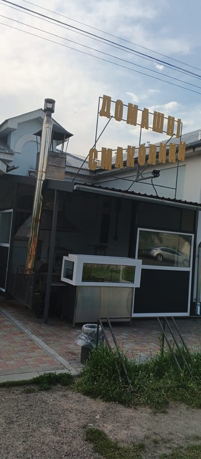 In Ploske, Ukraine, a yellow sign with Cyrillic lettering, reading "ДОМАШНІ СМАКОЛИКИ" (which translates to "homemade delicacies"), is mounted on a metal frame against a cloudy sky. Below the sign, a small open-air establishment is visible. It features a large, gleaming stainless steel chimney on the left, connected to some cooking equipment. To the right, there are large display windows, one of which reflects a silver car. The area in front of the establishment is paved with brick-like tiles and gravel. In the foreground, there is a trash can, some green grass, and a metal rack.
