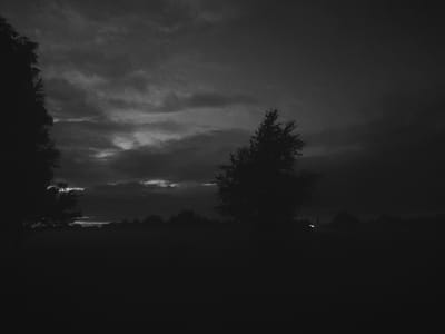 The image is a black and white photograph of a dark, cloudy sky over a silhouetted landscape. A lone tree with a full canopy stands in the middle ground, to the right of the frame. The foreground is a dark, featureless expanse, likely a field. On the far left, the silhouette of another tree or cluster of trees is visible. In the distance, along the horizon, a faint line of trees is discernible. A single, small, bright light source, possibly a distant lamp or structure, is visible in the far right mid-ground, barely breaking through the darkness. The overall mood is somber and overcast. The location is identified as Yaglavitski Sel'ski Savet, Belarus.