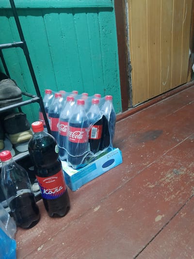 This media file shows a collection of Coca-Cola bottles and other items in what appears to be a storage area or hallway. The primary subjects are several plastic bottles of Coca-Cola, some in a multi-pack wrapped in plastic, and two individual bottles in the foreground. There's also a blue and white cardboard box with the word "INTEX" visible. In the background, a black metal shoe rack is visible with several pairs of shoes on it. A teal-colored wooden wall and a light brown wooden door are also present, suggesting an indoor setting.

The lighting suggests it might be daytime, possibly indoors with artificial lighting. There are no people visible in the image. The floor is made of worn wooden planks. The overall impression is one of storage or a hallway cluttered with beverages and shoes.

Visible text includes "Coca-Cola CLASSIC", "TƏKRAR EMAL ET", "КАРАЧИНСКИЙ Источник", "Экстра-", and "INTEX Krystal Clear Pool Basics". The Russian text indicates the origin of the beverages.