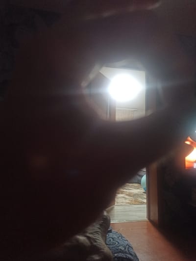 The media file shows a dimly lit interior scene, likely a room in a house or apartment. In the foreground, a blurred human hand or arm is visible, positioned as if holding a camera or phone. The focus is directed towards a bright, circular light source in the mid-ground, possibly a lamp or a screen emitting light. Behind this light, a white structure with vertical lines, perhaps a door or a part of the room's architecture, is faintly discernible. Further in the background, hints of a wooden floor and a patterned fabric, possibly a blanket or upholstery, can be seen. A portion of what appears to be a television screen displaying an image is visible in the upper right corner of the frame, emitting a warm orange glow. The overall impression is of a private, indoor space, possibly at night or with the lights dimmed. No specific activity is clearly depicted, beyond the implied act of taking a photograph. The lighting conditions suggest a low-light environment, with a strong light source creating lens flare and obscuring details. There is no visible text in the image. The provided location context of Rubtsovsk, Russia, does not offer any specific visual cues within the image itself.