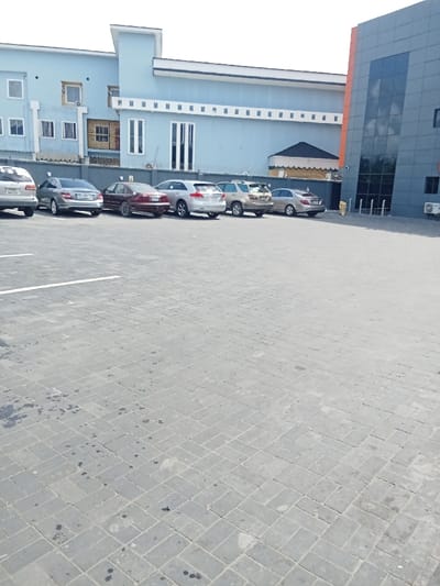 The image shows an empty parking lot with seven vehicles parked in a row. The parking lot is made of gray interlocking pavers and is in front of a two-story building with a light blue facade. There is a taller building to the right of the image. The taller building has a dark gray facade with large glass windows. The location, based on the provided coordinates, is Ujevwu, Nigeria.