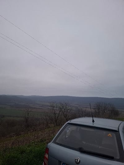 The scene depicts a rural landscape in Balkantsi, Bulgaria, under an overcast grey sky. The lower right portion of the frame shows the rear section of a light grey Skoda hatchback, positioned on a grassy verge. The vehicle is equipped with a roof-mounted antenna, a rear windshield wiper, a Skoda emblem on the tailgate, and a yellow triangular warning sticker affixed to the upper right corner of the rear window. Behind the vehicle, the terrain slopes downward, covered by green grass and sparse, leafless deciduous trees. The background features extensive rolling hills and valleys, exhibiting a mosaic of agricultural fields and wooded areas extending to the horizon. Four parallel power or communication lines traverse diagonally across the upper left section of the sky. No individuals are visible in the scene.