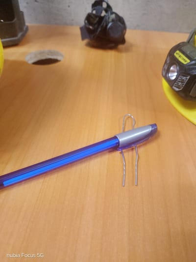 This image shows a close-up view of a wooden desk surface. The main subject is a blue pen with a silver tip, which is partially held up by a silver paperclip. The paperclip is bent in a way that creates a small stand for the pen.
In the background, several objects are visible, though they are out of focus. On the left, a dark, possibly plastic object is partially visible. In the middle background, there are two black objects that appear to be headlamps or flashlights, one of which has a yellow band. The surfaces appear to be made of wood and a textured gray material.
The lighting suggests an indoor setting, likely during the daytime, as there is no indication of artificial lighting sources. The overall scene is static, with no apparent activity taking place.
Visible text includes "NITECORE" on one of the headlamps and "nubia Focus 5G" in the bottom left corner of the image.