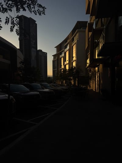 The image captures a street scene in Ciudad Santa Catarina, Mexico, taken at dusk. The street is lined with modern buildings on both sides. The building on the left is tall, featuring a billboard with a faded image and some writing. There is a line of parked cars on the left side of the street. A building on the right features an awning and appears to be a commercial property. There is a sidewalk with a row of chairs and a table between the buildings. The buildings appear to be constructed of concrete and glass. The scene is lit by the setting sun, casting long shadows across the street and buildings. The sky is a deep blue.