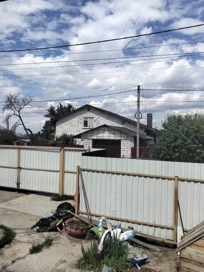 The image captures a scene in Sloboda, Russia, featuring a partially constructed house made of white cinder blocks. The house has a pitched roof, a single window with white blinds, and a garage door. In the foreground, a tall, white metal fence with wooden posts runs horizontally across the frame, partially obscuring a cluttered yard. This yard contains several black trash bags, a pink planter with soil and plants, a red traffic cone, a broom, and various pieces of debris including plastic items and a blue watering can. A bare, gnarled tree stands to the left of the house, and dense green foliage surrounds the property. Overhead, power lines crisscross the cloudy blue sky. The overall impression is one of unfinished construction and unkempt outdoor space.