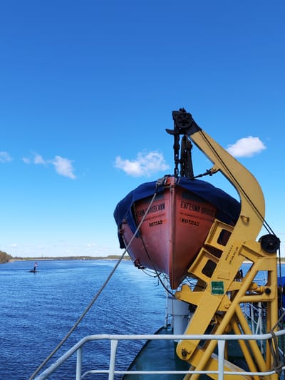 The image depicts a lifeboat on a yellow davit against a backdrop of a bright blue sky with scattered clouds and a river or wide waterway. The lifeboat is primarily orange-red with a dark blue cover partially obscuring its interior. The name "ЕВГЕНИЙ ОРЛОВСКИ" (Evgeniy Orlovskiy) is painted on the side of the boat in Cyrillic, along with text indicating "AO РЫБИНСКОЕ ПАРОХОДСТВО" (Rybinsk Shipping Company) and the city "ОСКОТЛАС" (Kotlas). Other markings on the boat, partially obscured, contain the name "АГЕНИЙ СЛОВСКИЙ" and "КОТЛАС." There are additional, smaller markings on the davit structure, and a warning sign with a symbol of a wave is visible.

The waterway has a buoy or navigation marker visible in the distance. The scene is likely taken from a vessel or dock, based on the railing and deck visible in the foreground. The geographical location is unknown, but the Cyrillic text suggests the image was taken in a Russian-speaking area. There is no indication the image was taken in Chaika, Russia.