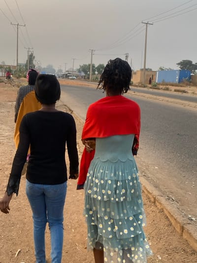 Three people stand on a roadside. The first wears a blue dress with white daisies and a red shawl. The second wears a black shirt and blue jeans. The third wears a yellow shirt. All three face away from the camera and toward the road. Power lines are visible in the background. A blue building is in the distance. The sky is hazy.