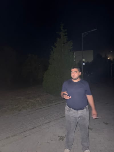 A medium shot captures a man standing on a paved walkway at night. He is wearing a dark blue t-shirt and light gray jeans. In his left hand, he holds a dark-colored smartphone, and in his right hand, he is holding a cigarette. The background features a tall evergreen tree to his left and a building in the distance. A streetlamp is visible further in the background. The ground is covered in interlocking pavers. The scene appears to be in an outdoor urban environment.