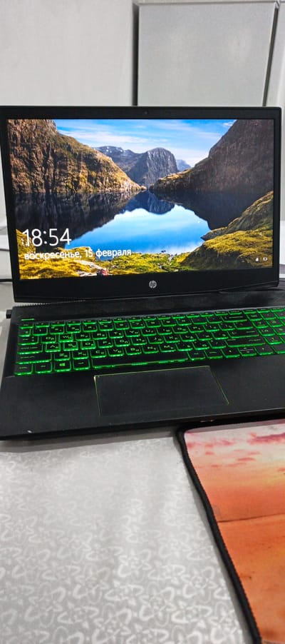 The scene is located indoors in Moynaq, Uzbekistan. A black HP laptop is positioned on a light-colored surface with a subtle, repeating floral pattern. The laptop screen displays an active desktop background or lock screen featuring a natural landscape with a calm lake reflecting rugged mountains and green terrain under a blue sky. Overlaid on this image are the white text "18:54" and "воскресенье, 15 февраля" (Sunday, February 15th). The laptop's QWERTY keyboard, which also includes Cyrillic characters, is illuminated with a bright green backlight. A portion of a mouse pad, featuring a sunset or sunrise image with warm orange and pink hues, is visible on the surface to the right of the laptop. The background consists of indistinct, light-colored wall sections. No individuals or active interactions are present in the image.