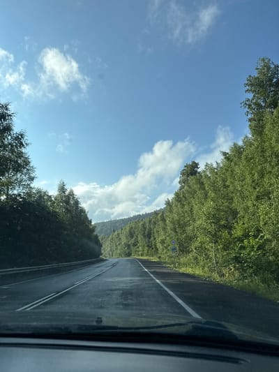 This image depicts a view from a vehicle driving on a wet, asphalt road, surrounded by dense green foliage. The road, marked with double white lines, curves gently into the distance, leading towards a backdrop of rolling hills covered in trees. The sky is a bright blue with scattered white clouds, illuminated by sunlight. On the right side of the road, there are a couple of road signs, one circular with the number "70" indicating a speed limit, and another rectangular sign below it. The trees lining the road are tall and appear to be a mix of deciduous and coniferous species. The overall scene suggests a journey through a forested area in a rural or natural setting. The location is identified as Brishbash, Russia.