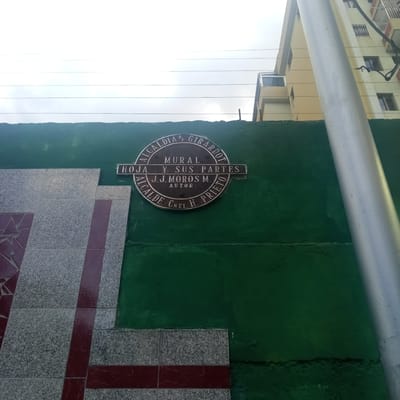 The image shows a circular plaque mounted on a green wall. The plaque is bronze and has text inscribed on it. The text reads "ALCALDIA DE GIRARDOT" around the top edge, "MURAL HOJA Y SUS PARTES" in the center, and "J.J. MOROS M. AUTOR" below that. Around the bottom edge of the plaque, it reads "ALCALDE CNEL. H. PRIETO". The plaque is located outdoors, with a building visible in the background. The building appears to be a light-colored, multi-story structure with windows and balconies. A silver pole is also visible in the foreground, running vertically alongside the plaque and wall. The green wall has a rough texture, and some parts appear to be peeling. To the left of the plaque, a section of a tiled wall is visible, featuring a pattern of gray speckled tiles and darker red stripes. The sky is overcast, with visible power lines stretching across the upper portion of the image. The scene depicts an outdoor public space, likely a wall or building facade, in El Limón, Venezuela, as indicated by the reference to "Alcaldia de Girardot", which is the municipality where El Limón is located.
