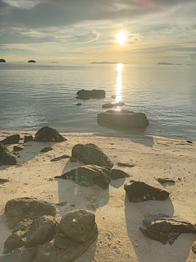 The image depicts a tranquil beach scene at Baan Pang Ka, Thailand, during sunset. The sun, a bright orb, casts a shimmering reflection on the calm sea, with a few islands dotting the horizon. The foreground is occupied by a sandy beach littered with dark, weathered rocks of various sizes. Some of these rocks are partially submerged in the gentle waves lapping at the shore. The sky is a gradient of soft hues, with scattered clouds illuminated by the setting sun. No people are visible in the image.