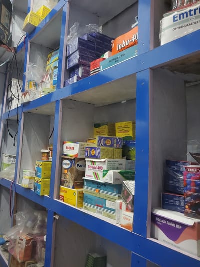 The image shows shelves in a pharmacy, likely in Mutum Biyu, Nigeria, filled with various boxes of medications. The shelves are painted blue and white, with some of the white surfaces showing signs of wear. On the shelves, different brands and types of medicine are visible, including boxes with labels like "Emtrim," "Inbu-40," "Norbactin 400," "Strovid-400," "Para1000," and "Ofloxacin Tablets USP 400mg." Some boxes are stacked in neat rows, while others are placed more haphazardly. Plastic packaging is evident on some of the medicines. Cables from electronic devices are also visible in the background, suggesting the presence of equipment within the pharmacy. The overall impression is of a working pharmacy, well-stocked with pharmaceuticals.