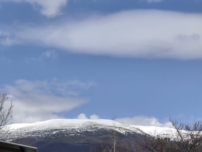 The image displays a wide, elevated view of a snow-covered mountain range in Pirdop, Bulgaria, under a partly cloudy sky. The upper two-thirds of the frame are dominated by a clear blue sky, traversed by multiple white cloud formations, including a prominent, elongated band of stratus clouds across the top. The mountain range, positioned in the midground, exhibits significant snow cover on its upper slopes and peaks, with darker, possibly forested or rocky terrain visible below the snowline. In the lower left corner, the light-colored eave of a building's roof is partially visible. The lower right foreground features numerous bare, dark tree branches. Partially obscured by these branches, a dark, skeletal metal structure is discernible against the mountain. No human or animal figures are present.