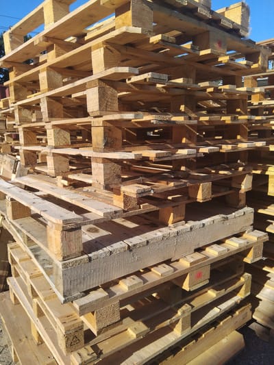 The image shows a large stack of wooden pallets outdoors. The pallets are of various sizes and conditions, with some appearing newer and lighter in color, while others are weathered and darker. They are stacked in a disorganized yet stable manner, reaching a significant height. The lighting suggests it is daytime, with shadows cast by the pallets. In the background, a clear blue sky is visible, with some green foliage peeking through from behind the pallets on the left side. A portion of a building's side, likely made of corrugated metal, is also visible on the far left. The scene appears to be an outdoor storage area, possibly a warehouse or industrial yard. The specific location of Deurne, Netherlands is not directly indicated by any visual cues in the image itself.