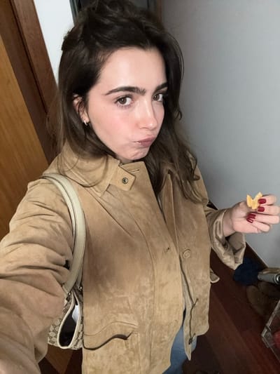 A woman poses in a selfie, holding a cracker. Her face is pale, with brown hair and eyes. She wears a tan suede jacket and a white purse with a patterned design. Her nails are painted dark red. The background shows a wall and wooden flooring with shoes placed on a wire rack. The image is slightly rotated.
