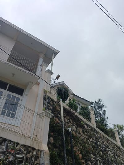 The image depicts an upward-angled view of a multi-level urban or semi-urban environment in Makilala, Philippines, under an overcast sky. In the foreground on the left, a light-colored, multi-story building features a second-floor balcony with white decorative columns and a white metal railing. A large white-framed window with a grid pattern is visible on the first floor, protected by a white metal fence. The building's lower section is supported by a dark, irregular stone foundation.
Centrally, a tall, multi-toned stone retaining wall, heavily covered with green vines and vegetation, rises diagonally from left to right. This wall is topped with a light-colored concrete coping and a dark metal fence, possibly incorporating razor wire. Two vertical utility pipes, one dark and one lighter, are affixed to the retaining wall.
Behind the retaining wall and on a higher elevation, a second, lighter-colored building is partially visible. It features a dark-tiled or shingled gabled roof and white-framed windows, with a dark metal railing along its perimeter. A dark-colored street lamp with a horizontal fixture is positioned above the retaining wall and in front of the second building.
In the upper right quadrant, two parallel dark utility lines traverse the uniformly gray, overcast sky. Various green shrubs and tall, slender-leafed plants are present on the elevated ground behind the retaining wall, adjacent to the second building. No individuals or dynamic actions are depicted.