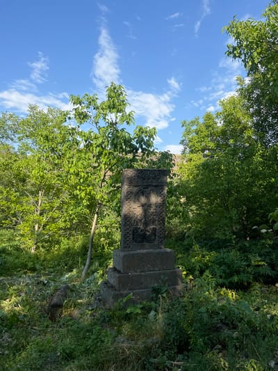 An ornate stone cross stands prominently among lush greenery, under a sky of soft blue and scattered clouds. The cross's intricate carvings contrast with the surrounding foliage. The structure is comprised of multiple stacked stone pieces. The verdant background enhances the cross's aged texture. Sunlight dapples through the canopy above, casting shadows on the historical monument. The scene evokes a sense of timeless beauty and serenity.