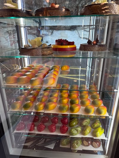 A display case features an array of desserts. Round chocolate cakes with varying decorations are positioned on the left. Multiple rows of fruit-shaped treats, including mangoes, green fruits, and raspberries, are neatly arranged on stands. Vanilla beans are visible to the right. The glass case reflects an outdoor scene, partially obscuring the desserts.