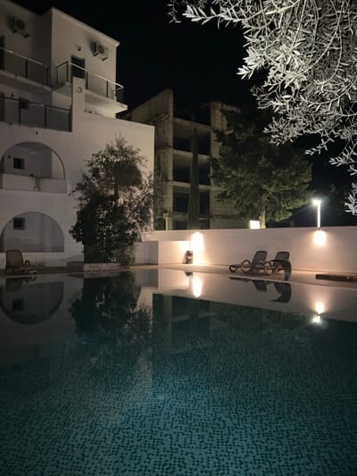 The image shows a night view in Brežine, Montenegro. A multi-story building with white walls, balconies, and air conditioning units is on the lower left. Above it and slightly to the right is a taller, unfinished concrete building with multiple window openings. A tree with sparse, light-colored foliage is in the upper left corner of the frame. In the center, a brightly lit structure reflects in the water of what seems to be a pool. A tree is also reflected in the pool’s surface. The pool's tiles are visible. Overhead lights illuminate the central structure. A partially visible sign is present on the lit structure.