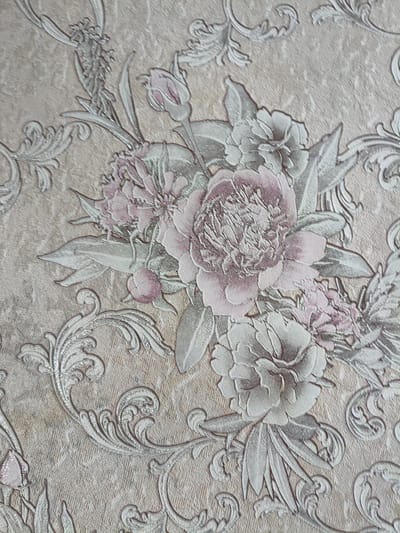 This image is a close-up of textured wallpaper featuring an embossed floral pattern. The wallpaper has a beige background with raised floral designs in shades of pale pink, white, and silver. The pattern includes large, detailed flowers that resemble peonies, with numerous petals, as well as smaller buds and decorative scrollwork. The texture of the wallpaper appears to be woven or fabric-like, with a subtle sheen from the silver accents. The lighting suggests a natural light source, casting soft shadows that highlight the embossed details of the floral elements and the background texture. There are no people or other objects visible in the image, and the scene is static. The location in Rîșcani, Moldova, is not discernible from the visual content of the image itself, as it depicts a generic interior design element.
