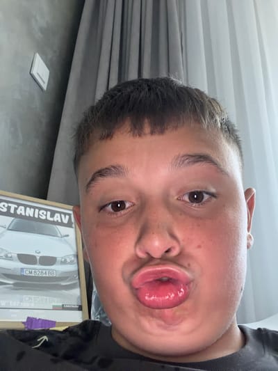 The image is a close-up, selfie-style photograph of a young person with short, dark hair and a fair complexion. The person is making a duck face, with their lips pursed. They have brown eyes and light eyebrows. Freckles are visible on their cheeks.

In the background, to the left, there is a framed picture of a silver BMW car with a Bulgarian license plate that reads "CM 8284 BB". The name "STANISLAV" is visible at the top of the frame. The picture is propped up against a grey wall with a white light switch.

To the right of the person, a grey curtain is visible, partially obscuring a white wall or window. The overall setting appears to be indoors, possibly a bedroom or living area. There is no information in the image to suggest a specific location within the city of Zmeitsa, Bulgaria.
