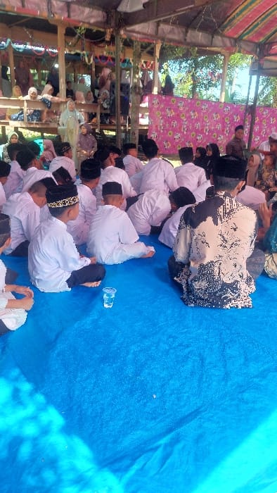 The scene depicts a communal gathering under a temporary open-sided structure in Jambo Dalam, Indonesia. A large group of approximately two dozen young boys, predominantly dressed in white long-sleeved shirts and black peci (traditional caps), are seated on a bright blue tarp covering the ground in the foreground and mid-ground, facing away from the camera. One boy in the immediate foreground wears a black peci with a golden patterned band. An adult male, visible from behind, sits among the children, wearing a patterned long-sleeved shirt and a dark peci. A clear plastic cup containing liquid rests on the blue tarp in the lower-left. In the background, an elevated section constructed of wooden planks hosts several women, most wearing headscarves, observing the event. The temporary structure features exposed wooden support beams and a roof, with colorful decorative fabrics and a pink patterned backdrop visible in the background. The environment suggests an outdoor event under partial shelter during daylight.