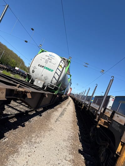 The image shows a train on tracks, likely in Dudelange, Luxembourg. A large white tank container with a green frame, labeled "GCA CONTAINER," sits on a flatcar. The tank bears the identification number GCAU 798068 8 EMKD. Details such as maximum gross weight (39000 KG / 85980 LB) and tare weight (4200 KG / 9259 LB) are visible. Above the train, overhead electrical lines are strung with numbered markers (04, 05, 06). The railway tracks run along a gravel bed. Other train cars are visible behind the tank container. A small patch of trees is visible in the distance. The sky is clear and blue.