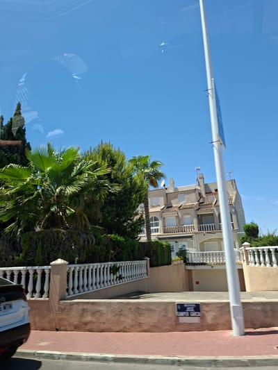 The image shows a residential street in La Mata, Spain, with a row of three-story townhouses. The houses are painted a light beige color with white trim and feature balconies with white railings. There are palm trees and other lush greenery in front of the houses. A white street sign is visible on a low wall in the foreground, reading "RONDA JOSE SAMPER GARCIA". A white car is parked on the side of the street. The sky is a clear blue, indicating a sunny day. The scene is viewed from a slightly elevated perspective, suggesting the photo may have been taken from a vehicle.