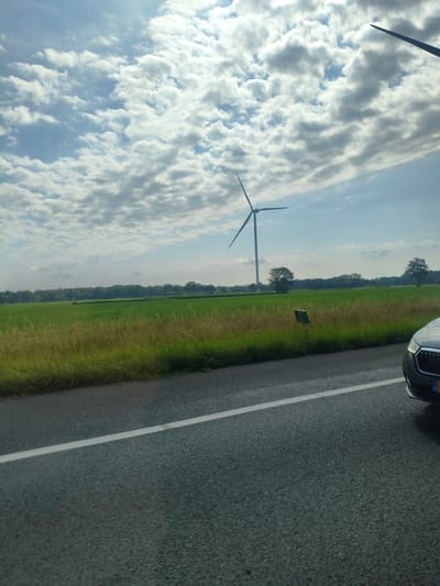 The image depicts a rural landscape under a cloudy sky, likely in the outskirts of Breda, Netherlands. A dark-colored car is partially visible in the foreground, driving on an asphalt road with a white lane marking. Beyond the road, a green field stretches towards a treeline. In the middle ground, a wind turbine stands tall, its blades extended against the sky. The overall scene conveys a sense of open space and natural elements, possibly captured during a journey through the Dutch countryside.
