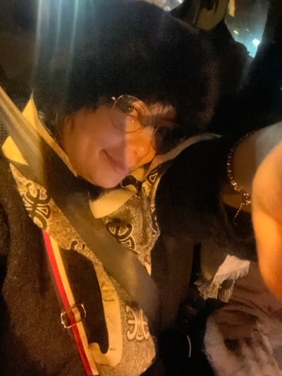 A person with round glasses is shown in an upward-angled shot. They have dark hair and are wearing a black top. A chain necklace adorns their neck. A patterned fabric lies near their face, and a grey strap with a red, white, and cream band is visible. The overall lighting is dim, with dark and light areas.