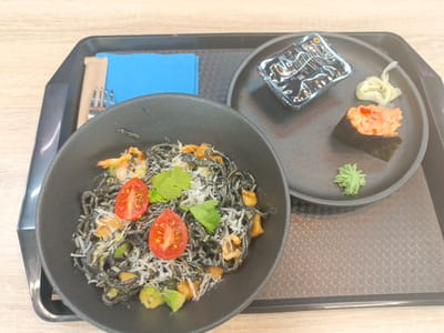 The image shows a meal on a black tray. On the left, a black bowl contains dark spaghetti with tomatoes, herbs, shredded cheese, and avocado. Next to the bowl, there's a blue napkin with a fork and a knife. On the right, a black plate holds a container of sauce, a piece of sushi, and two small mounds of garnishes (ginger and wasabi). The tray sits on a wooden surface. The location is Novoivanovskoe, Russia.